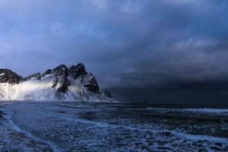 White foamy waves on the beach of Stokkness, behind snowy rocky slopes of Vestrahorn, near Höfn,