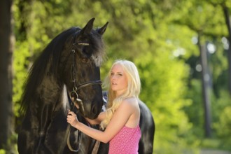 Blonde Woman in a pink dress holding a black Friesian horse on its bridle in a forest setting,