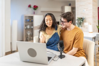 Multiethnic young couple using a laptop and a microphone, recording a podcast from their home