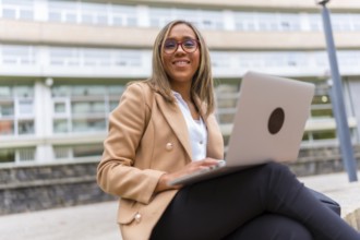 Happy latin woman entrepreneur smiling at camera using laptop sitting in the city
