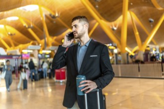 Businessman making a phone call while holding a reusable coffee cup and pulling his luggage at a