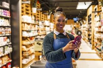 Smiling saleswoman using a smartphone in an organic supermarket with shelves full of natural and