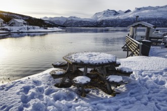 Snow-covered table and benches by the lake with mountains in the background, quiet winter