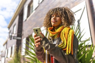 Latin young woman using phone standing in a residential street wearing warm clothes and scarf in a
