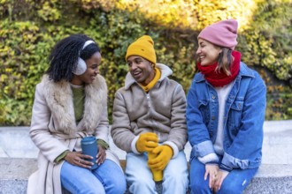 Three cheerful young friends are sitting on a bench in a city park during winter, holding reusable