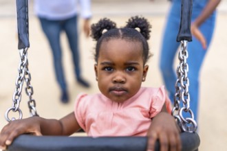 Cute african baby girl sitting on swing in a public park next to her family