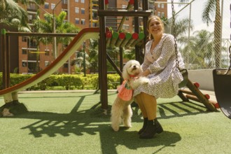Happy woman sitting on a swing in a vibrant playground, joyfully embracing her small white dog