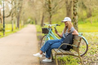 Blonde female tourist wearing casual clothing, using smartphone and resting on park bench with