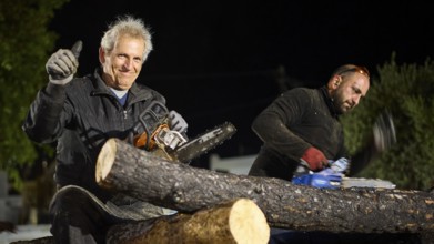 ©Udo Rein, An elderly man gives a thumbs up while sitting next to a woodworker with a chainsaw,