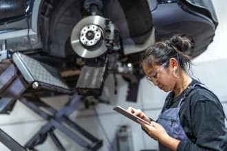 Young female mechanic using a digital tablet while inspecting a car raised on a hydraulic lift in a