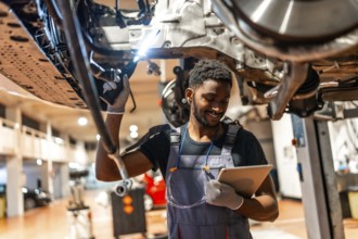 Young mechanic using a digital tablet while working on a car held up by a hydraulic lift in a