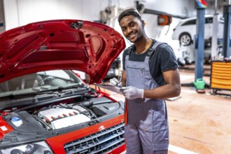Smiling mechanic taking notes on clipboard while inspecting engine of red car with open hood in a