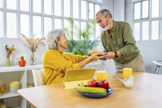 Senior man serving breakfast to share with his wife while she is using digital tablet sitting on
