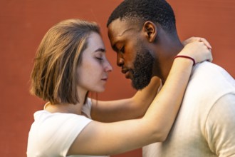 Multi-ethnic couple showing affection and hugging each other against red urban wall