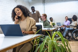 Woman talking to the mobile phone and using laptop in a coworking space with coworkers on the