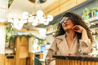 Elegant latin beauty woman with curly hair waiting hand on chin in a modern cafeteria