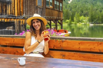 Young woman wearing straw hat smiling and holding a mug in a restaurant by lake dobbiaco with a