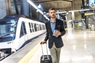 Businessman walking in subway station pulling suitcase and holding smartphone and reusable coffee