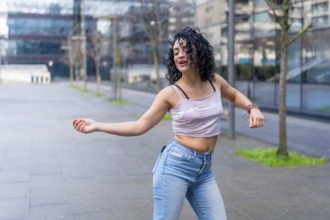 Curly haired hispanic woman dancing in urban city street with modern buildings in the background,