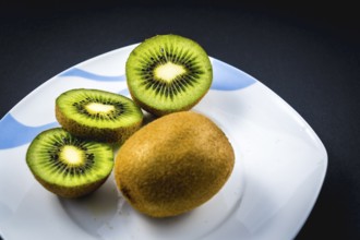 One whole and two cut kiwi on a white plate with a black background