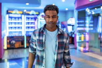 Young man smiles while standing in a lively arcade, surrounded by colorful claw machines and other