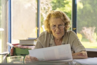 Focused elderly woman with glasses reviewing paperwork, possibly bills or medical records, sitting
