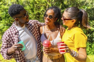Three young multi ethnic friends enjoying a sunny day outdoors, holding drinks and smiling