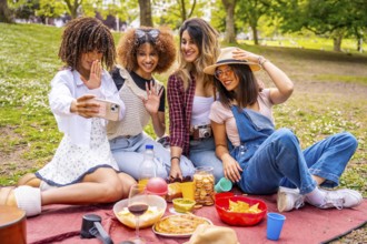 Four cheerful young women enjoying a picnic in a park, taking a joyful selfie with a smartphone