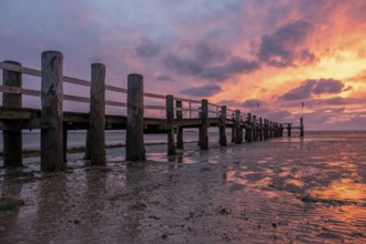 A jetty on the beach at sunset, the sky is coloured orange and purple, the water reflects the