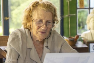 Elderly woman wearing glasses is sitting at a table, carefully reading documents with a focused