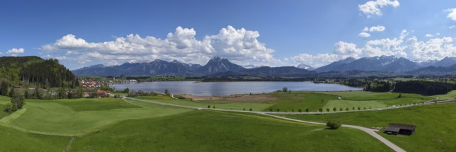 Expansive landscape with a view of Lake Hopfensee and mountains, surrounded by green meadows and a