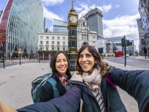 Two happy tourists taking a selfie in london near a historical clock tower, with modern buildings