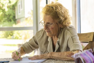 Concentrated elderly woman wearing glasses drawing with green colored pencil on sheet of paper