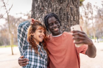 Young multiracial couple embracing and taking a selfie with a smartphone in a park, enjoying a