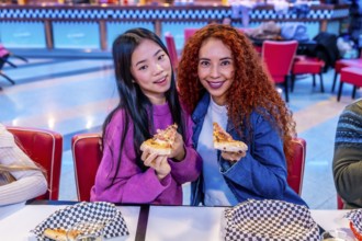 Two young women enjoying pizza slices at a bowling alley restaurant, celebrating friendship and