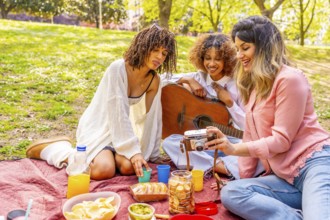Three female friends enjoying a picnic in a park, taking pictures with a vintage camera, eating