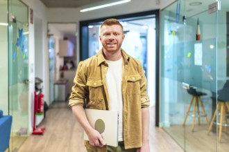 Young professional walking in a modern office corridor, holding a laptop and smiling, showcasing