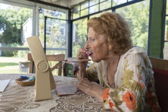 Senior woman sitting at a table in her cozy home, applying red lipstick with a small mirror,