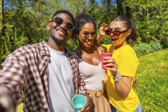 Three friends are taking a selfie while enjoying drinks outdoors, capturing a fun moment together