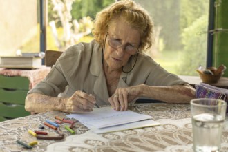 Elderly woman wearing glasses concentrating on drawing with crayons at a table with a glass of