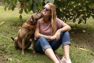 Woman in her 40s wearing sunglasses with a joyful expression receives a lick from her pet while