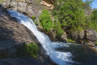 A small waterfall flows over large rocks, surrounded by green plants under a sunny sky, Cascada del