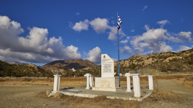 White memorial with Greek flag against a clear blue sky and mountain backdrop, Christopher Litos