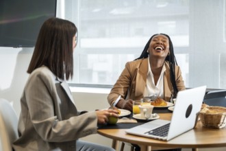 Two businesswomen are enjoying breakfast and laughing together during their business trip, creating
