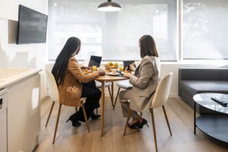 Two businesswomen are enjoying breakfast and working with laptop and tablet in a modern hotel room
