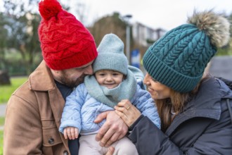 Parents wearing winter hats embracing their smiling baby in a park, enjoying a sunny winter day