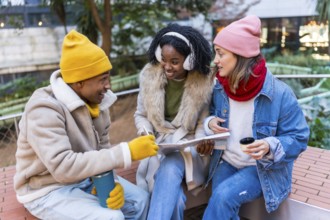 Three happy friends sitting outside in winter, drinking coffee and looking at a map, planning a