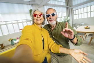 Senior woman with heart shape sunglasses taking selfie with husband while having fun at home