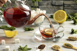 Red tea is poured from a pot into a glass cup with lemon and mint, surrounded by sugar and teas