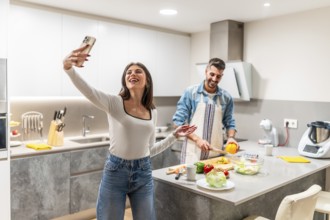 Happy young couple taking selfie with smartphone while preparing healthy food in modern kitchen